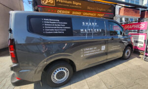 A side-profile view of a dark grey commercial van featuring custom professional vehicle graphics for "Sharp Matters." The design is clean and high-contrast, utilizing white text on the dark vehicle body and window tint: Main Branding: The "SHARP MATTERS" logo is prominently displayed on the side window, featuring a stylized hashtag-style graphic. Service List: A bulleted list on the rear window section details services: Knife Sharpening, Knife Repairs & Restoration, Scissors & Garden Tools, Commercial Kitchen Contracts, Domestic Appointments, and Custom Knife Making. Contact Information: The lower door panel includes a phone number (07949 892 193), website (sharpmatters.co.uk), email address, and a scannable QR code. Tagline: To the right of the contact details, the text "Trusted by chefs, loved by home cooks" is applied in a stylized script.