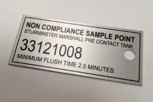 A rectangular metal tag with a single mounting hole on the right side, used for facility water testing. The black-etched text includes: Heading: "NON COMPLIANCE SAMPLE POINT". Location: "STURMINSTER MARSHALL PRE CONTACT TANK". ID Number: A large, bold "33121008". Instruction: "MINIMUM FLUSH TIME 2.5 MINUTES".