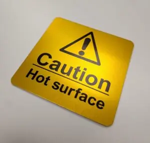 A square, metallic yellow safety sign with rounded corners and a brushed texture. The label features high-contrast black text and iconography. The sign contains: Icon: A black equilateral triangle containing a bold exclamation point, centered at the top. Text: The words "Caution" and "Hot surface" stacked vertically in a bold, sans-serif font. Detail: A horizontal line separates the word "Caution" from "Hot surface".