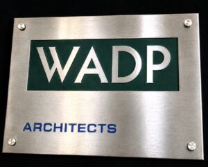 A large, rectangular stainless steel plaque with a brushed vertical grain. It features a modern architectural branding layout: Logo: The acronym "WADP" is knock-out etched in silver against a solid sage-green rectangular background. Secondary Text: The word "ARCHITECTS" is printed below in a bold, blue sans-serif font. Mounting: Four small pre-drilled holes are located in each corner for flush mounting.